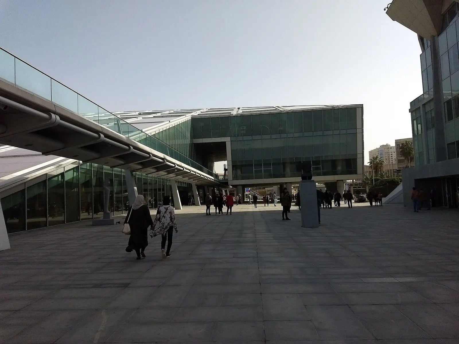 Visitors crossing the plaza in front of the glass facade of the Bibliotheca Alexandrina